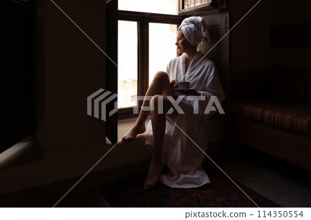 Woman in dark room sitting in robe by window. Attractive young girl in bathrobe and with towel on her head is holding cup, looking at window and smiling while sitting on window sill in old hotel room Woman in dark room sitting in robe by window. Attractive young girl in bathrobe and with towel on her head is holding cup, looking at window and smiling while sitting on window sill in old hotel room 114350554