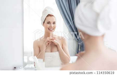 Woman wrapped in towel is examining her face in mirror, focusing on her skin, lips, eyebrows. She smiles at her reflection. Girl with towel on head takes care of her facial skin Woman wrapped in towel is examining her face in mirror, focusing on her skin, lips, eyebrows. She smiles at her reflection. Girl with towel on head takes care of her facial skin 114350619