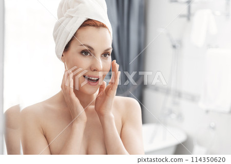 Woman wrapped in towel is examining her face in mirror, focusing on her skin, lips, eyebrows. She smiles at her reflection. Girl with towel on head takes care of her facial skin 114350620