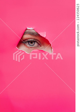 Eye of the beautiful woman looking through hole in pink paper.  114350623