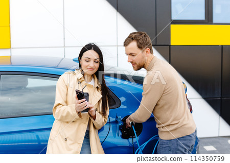 Young couple man and woman traveling by electric car having stop at charging station. Boyfriend plugging in cable to charge. Man talking with girlfriend, holding cup drinking hot coffee smiling Young couple man and woman traveling by electric car having stop at charging station. Boyfriend plugging in cable to charge. Man talking with girlfriend, holding cup drinking hot coffee smiling 114350759