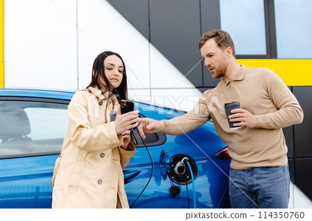 Young couple man and woman traveling by electric car having stop at charging station. Boyfriend plugging in cable to charge. Man talking with girlfriend, holding cup drinking hot coffee smiling Young couple man and woman traveling by electric car having stop at charging station. Boyfriend plugging in cable to charge. Man talking with girlfriend, holding cup drinking hot coffee smiling 114350760