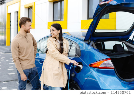 Young couple man and woman traveling by electric car having stop at charging station. Girlfriend holding charger looking at car and smiling Young couple man and woman traveling by electric car having stop at charging station. Girlfriend holding charger looking at car and smiling 114350761