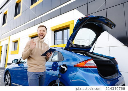 A young man is charging an electric car while standing at a station and holding a tablet A young man is charging an electric car while standing at a station and holding a tablet 114350766