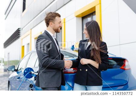Smiling woman standing at the car and pointing at car seller tablet. She is picked right car for her she saw online. A woman concludes an agreement to buy a car and shakes the manager's hand Smiling woman standing at the car and pointing at car seller tablet. She is picked right car for her she saw online. A woman concludes an agreement to buy a car and shakes the manager's hand 114350772