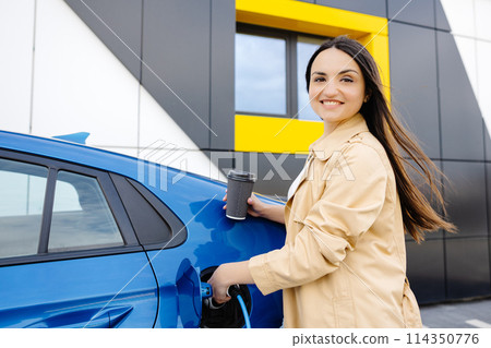Happy young woman charging automobile battery from small public station, standing near electric car, drinking coffee Happy young woman charging automobile battery from small public station, standing near electric car, drinking coffee 114350776