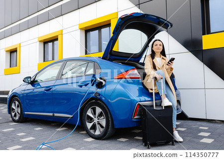 Young woman waiting charging automobile battery from small public station and using smartphone while charging automobile Young woman waiting charging automobile battery from small public station and using smartphone while charging automobile 114350777