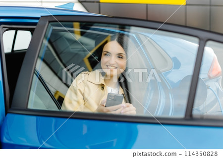 Business lady sitting inside charging electric car and using modern smartphone. Technology and transport concept. Business lady sitting inside charging electric car and using modern smartphone. Technology and transport concept. 114350828