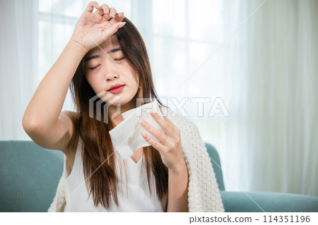 Sick woman sitting under blanket on sofa and sneeze with tissue paper in living room, Asian young female blowing nose sneezing in tissue at home, fever caught cold Sick woman sitting under blanket on sofa and sneeze with tissue paper in living room, Asian young female blowing nose sneezing in tissue at home, fever caught cold 114351196