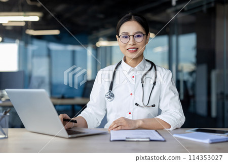 Professional and friendly female doctor wearing glasses and a stethoscope using a laptop in a contemporary office setting. Her confident smile suggests a high level of expertise and approachability. 114353027