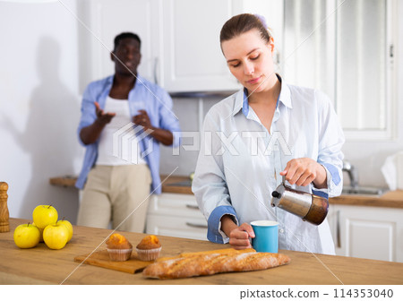 Upset young woman pouring coffee into cup on background of dissatisfied husband 114353040