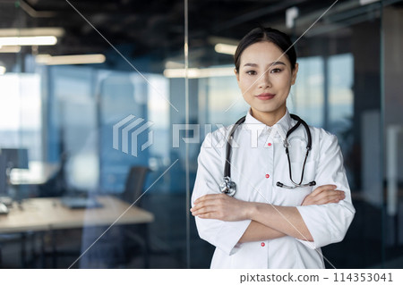 A serious Asian female doctor with crossed arms standing confidently in a modern hospital environment, showcasing professionalism and readiness. 114353041