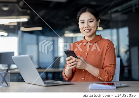 A professional businesswoman smiles while using her smartphone at a modern office workspace, exuding confidence and efficiency. 114353050