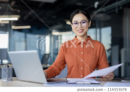 Confident businesswoman in glasses smiles while holding documents, working in a dynamic office setting with laptop open beside her. 114353076
