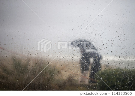 portrait of people walking on border the sea with umbrella behind a window with rain drops 114353500