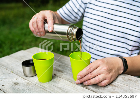 A man on the street pours coffee from a thermos into a green cup, enjoying his outdoor recreation. 114355697