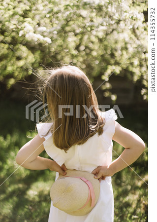 Portrait of little toddler girl in white romantic dress in blossoming garden in spring. Cute beautiful lovely child having fun in a park on a sunny day. 114355732