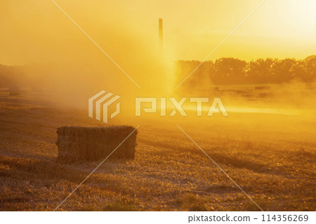 Combine harvester pressing straw from field into bales driving field on sunny summer evening. Field with bales of pressed wheat. Lots of dust on field. Agricultural agro industrial harvesting works Combine harvester pressing straw from field into bales driving field on sunny summer evening. Field with bales of pressed wheat. Lots of dust on field. Agricultural agro industrial harvesting works 114356269