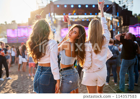 Group of young women enjoy summer music festival on sandy beach, holding drinks, dancing with stage lights behind. Casual outdoor party, sunset vibes. Fun at beachfront concert. 114357583