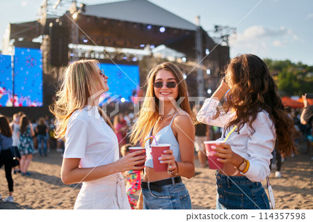 Group of joyful women with cool summery drinks at beach music festival. They dance under clear sky, stage with live performance in background. Girls in sunglasses enjoy fest vibes, sunlit ambiance. Group of joyful women with cool summery drinks at beach music festival. They dance under clear sky, stage with live performance in background. Girls in sunglasses enjoy fest vibes, sunlit ambiance. 114357598