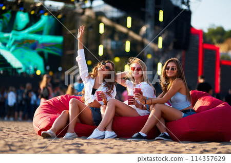 Three women sit on red bean bag chairs at beach music festival, toasting drinks, enjoying sun, with stage lights and crowd in background. Friends smile and wave, immersed in coastal event vibes. Three women sit on red bean bag chairs at beach music festival, toasting drinks, enjoying sun, with stage lights and crowd in background. Friends smile and wave, immersed in coastal event vibes. 114357629
