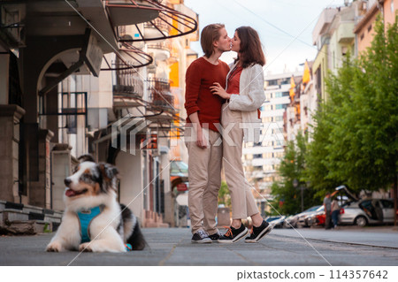 Young couple, man and woman kissing at the street. Dog at foreground. Family walking together in city with pet Young couple, man and woman kissing at the street. Dog at foreground. Family walking together in city with pet 114357642