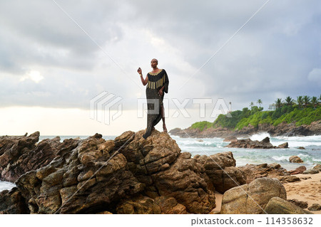 Gender fluid black person poses gracefully standing on rocks in ocean . Androgynous ethnic fashion model in posh dress and jewellery on rocky beach by storm. Pride month Gender fluid black person poses gracefully standing on rocks in ocean . Androgynous ethnic fashion model in posh dress and jewellery on rocky beach by storm. Pride month 114358632