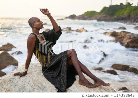 Non-binary black person in luxury dress, golden jewelry on beach rocks in ocean. Trans ethnic fashion model wearing jewellery in posh gown poses gracefully in tropical seaside location on sunset. Non-binary black person in luxury dress, golden jewelry on beach rocks in ocean. Trans ethnic fashion model wearing jewellery in posh gown poses gracefully in tropical seaside location on sunset. 114358642