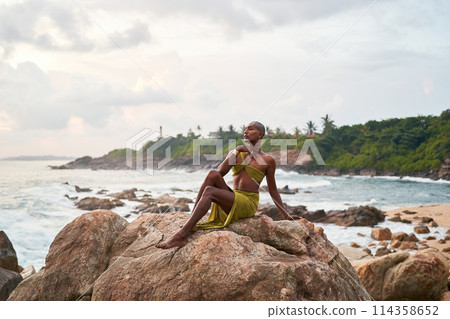 Androgynous black fashion model poses on scenic ocean beach at sunset. Non-binary ethnic fashion model in long revealing dress sits on rocks by sea against sunrise. Dusk on ocean background. LGBTQ 114358652