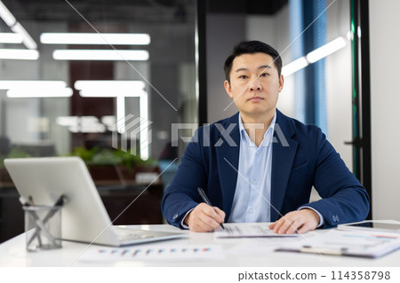 Focused Asian businessman in a well-fitted blue suit working at his office desk surrounded by documents and a laptop, exuding professionalism and determination. 114358798