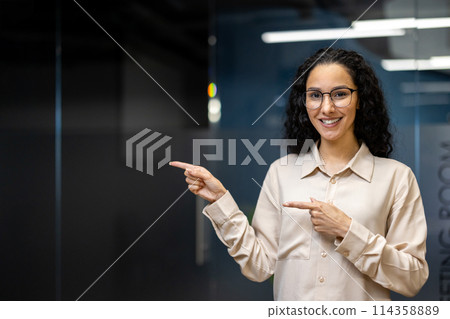 A Hispanic businesswoman in a beige shirt smiling and pointing to her side in a modern office setting, exuding confidence and professionalism. A Hispanic businesswoman in a beige shirt smiling and pointing to her side in a modern office setting, exuding confidence and professionalism. 114358889
