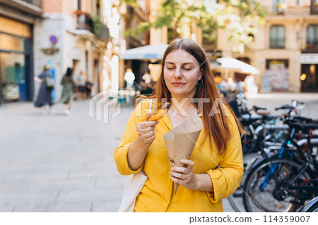 Carefree woman eating traditional Churros, a fried pastry with chocolate on a city street. Positive cheerful female student posing outdoors. Food concept 114359007