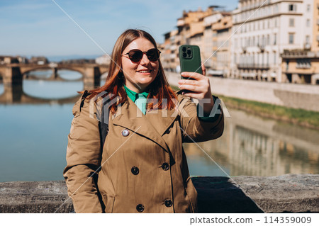 Attractive young female tourist is exploring new city. Redhead girl holding a smartphone on famous Old bridge in Florence. Traveling. Happy optimistic girl walking in city and makes selfie Attractive young female tourist is exploring new city. Redhead girl holding a smartphone on famous Old bridge in Florence. Traveling. Happy optimistic girl walking in city and makes selfie 114359009