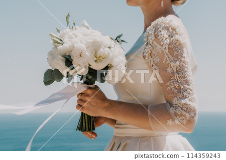 bride is holding a bouquet of white flowers. The bouquet is large and has a lot of flowers in it. The woman is wearing a white dress and a gold ring on her finger. Scene is elegant and romantic. 114359243