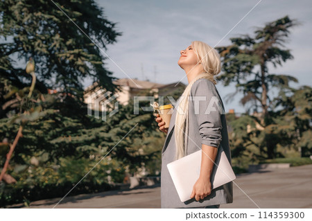 A woman is walking down the street with a white laptop bag and a cup of coffee. She is smiling and enjoying the sunny day. 114359300