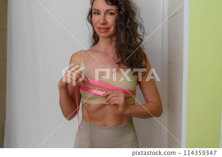 Cropped view of slim woman measuring breasts with tape measure at home, close up. Unrecognizable European woman checks the result of a weight loss diet or liposuction indoors. Healthy lifestyle. Cropped view of slim woman measuring breasts with tape measure at home, close up. Unrecognizable European woman checks the result of a weight loss diet or liposuction indoors. Healthy lifestyle. 114359347