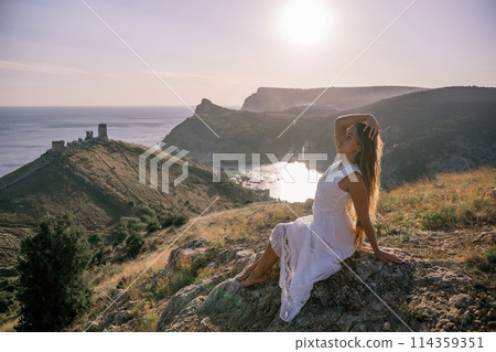 A woman in a white dress is sitting on a rock overlooking a body of water. She is enjoying the view and taking in the scenery. 114359351