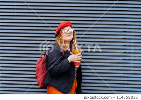 Smiling plus size woman in heart shaped sunglasses and bright clothes drinking sugar flavored tapioca bubble tea near striped urban wall. Happy fashionable hipster overweight girl. Street fashion. Smiling plus size woman in heart shaped sunglasses and bright clothes drinking sugar flavored tapioca bubble tea near striped urban wall. Happy fashionable hipster overweight girl. Street fashion. 114359818