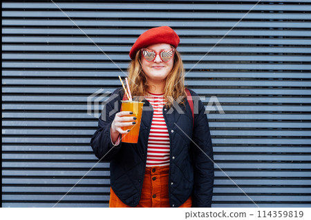 Smiling plus size woman in heart shaped sunglasses and bright clothes drinking sugar flavored tapioca bubble tea near striped urban wall. Happy fashionable hipster overweight girl. Street fashion. Smiling plus size woman in heart shaped sunglasses and bright clothes drinking sugar flavored tapioca bubble tea near striped urban wall. Happy fashionable hipster overweight girl. Street fashion. 114359819