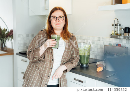 Smiling plus size young woman with just made glass of detox shake, green smoothie in the kitchen. Healthy dieting, eating, cooking, slimming. Natural weight loss program. Vegan, vegetarian diet. Smiling plus size young woman with just made glass of detox shake, green smoothie in the kitchen. Healthy dieting, eating, cooking, slimming. Natural weight loss program. Vegan, vegetarian diet. 114359832