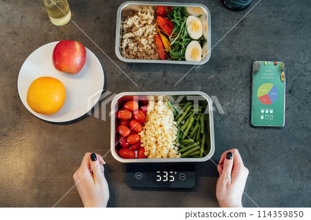 Healthy diet plan for weight loss, daily ready meal menu. Woman using meal tracker app on phone while weighing lunch box cooked in advance on kitchen scale. Balanced portion with dish. Pre-cooking 114359850