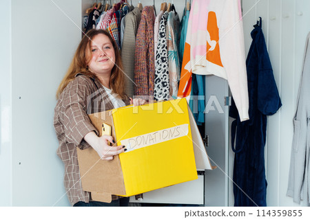 Smiling Woman with box of selected clothes from her wardrobe for donating to a Charity. Decluttering, Sorting clothes And Cleaning Up. Reuse, second-hand concept. Conscious consumer, sustainability 114359855