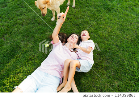 Cheerful cute boy kid having fun with his mom, embracing, laughing on the green grass with pet dog in garden on sunset. Summer outdoor activity. Happy active childhood, family time. Selective focus 114359865