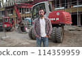 Portrait of successful builder, worker, contractor wearing hard hat and protective earphones, standing on work site with crossed arms confidently. Heavy construction equipment in background 114359959