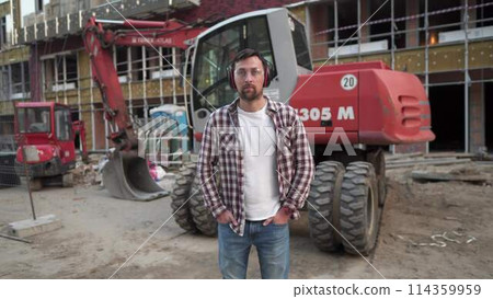 Portrait of successful builder, worker, contractor wearing hard hat and protective earphones, standing on work site with crossed arms confidently. Heavy construction equipment in background 114359959