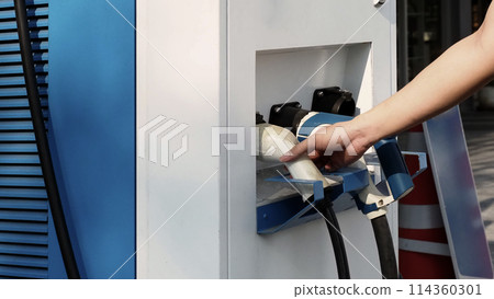 Driver of electric car hold batteries charger plug. Hand holding EV charging plug in front of camera with blurred background of outdoor. Sustainable energy powered electric charging station background Driver of electric car hold batteries charger plug. Hand holding EV charging plug in front of camera with blurred background of outdoor. Sustainable energy powered electric charging station background 114360301