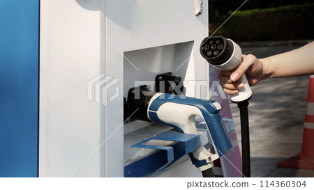 Driver of electric car hold batteries charger plug. Hand holding EV charging plug in front of camera with blurred background of outdoor. Sustainable energy powered electric charging station background 114360304