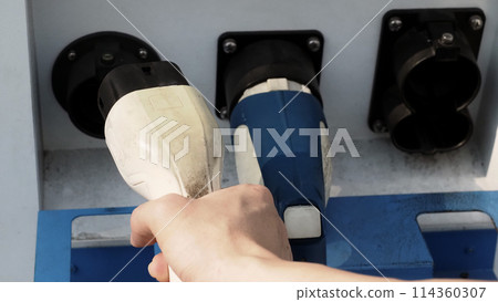 Driver of electric car hold batteries charger plug. Hand holding EV charging plug in front of camera with blurred background of outdoor. Sustainable energy powered electric charging station background 114360307