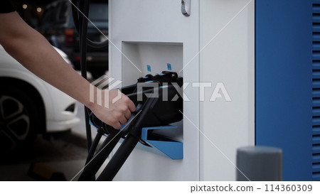Driver of electric car hold batteries charger plug. Hand holding EV charging plug in front of camera with blurred background of outdoor. Sustainable energy powered electric charging station background 114360309