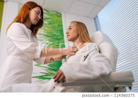Shooting from below of professional female cardiologist attaching holter sensors on woman patient with heart disease. Young woman during functional rehabilitation in cardiology room in clinic. 114360375